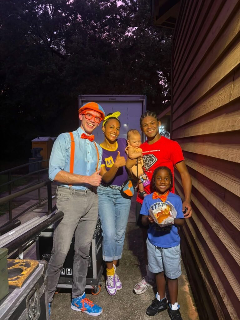 Two performers in Blippi-themed costumes posing alongside a local family including a man holding a baby, and a young child holding a food basket standing outdoors near a wooden wall and equipment cases.