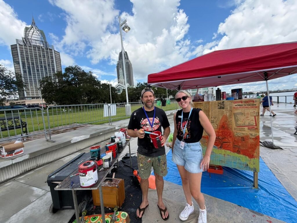 Two festival participants stand smiling next to an art supply table and a textured mural panel under a red canopy tent, with the downtown Mobile skyline in the background.