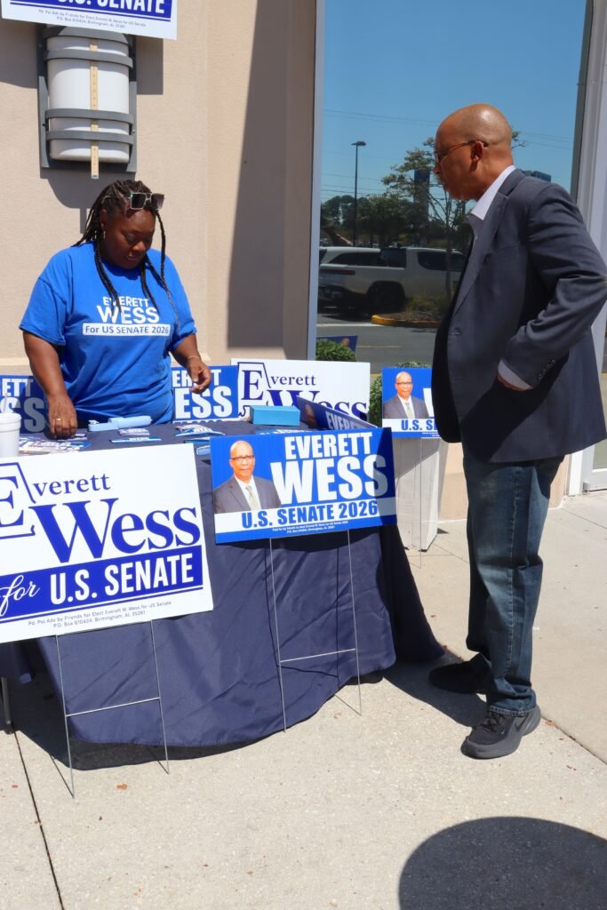 U.S. Senate candidate Everett W. Wess wearing a blazer and jeans standing outside next to a campaign volunteer at a decorated table.