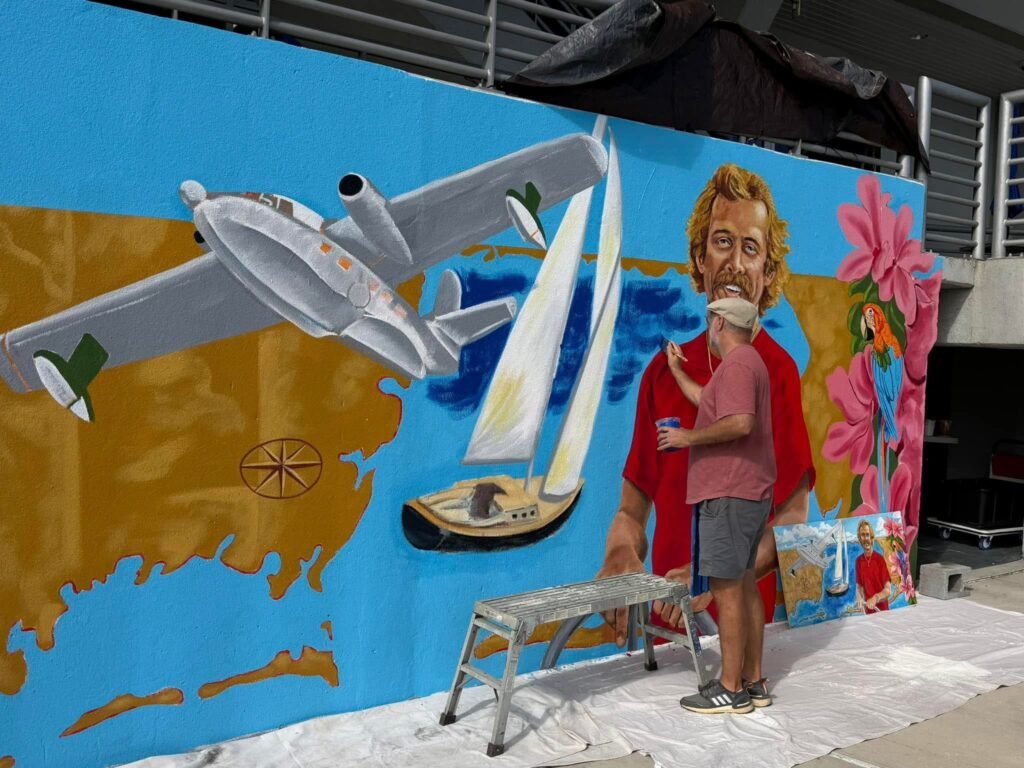 A close-up of an artist adding paint to a large outdoor mural depicting a vintage airplane, a sailboat on blue water, and a portrait of a man.