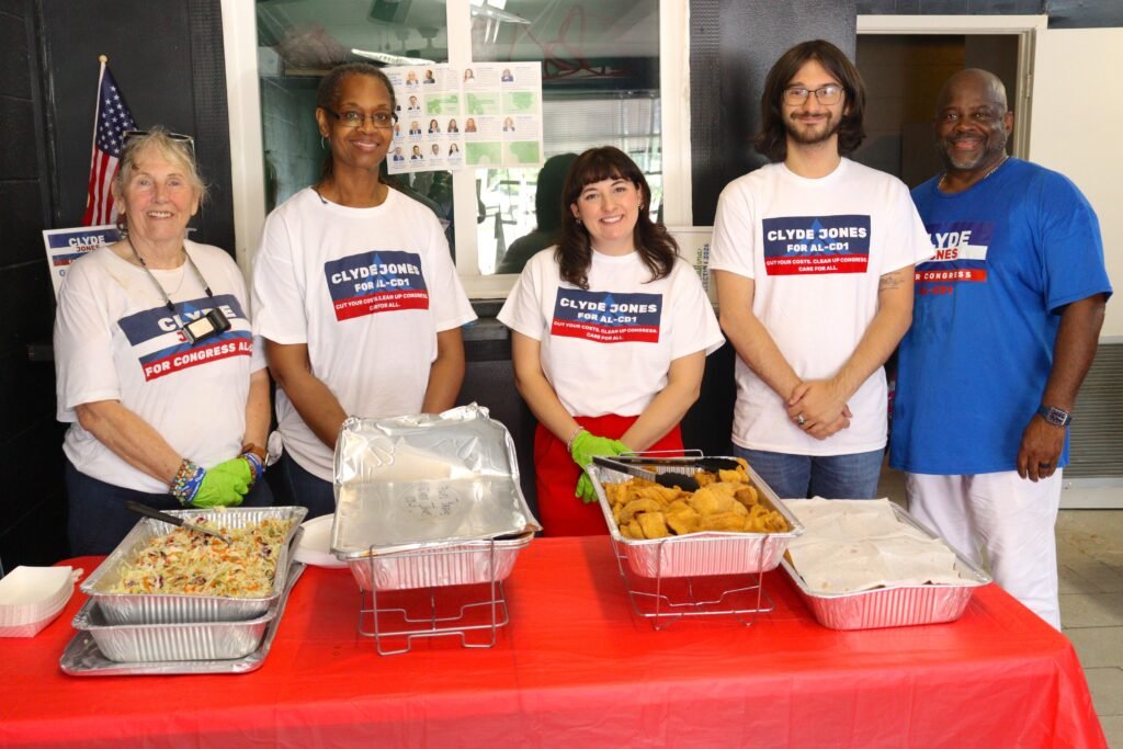 Four campaign volunteers, including Communications Director Payton Moran in the center wearing green serving gloves, stand behind a red table filled with catering trays of fried fish and coleslaw at the Mobile headquarters grand opening event.