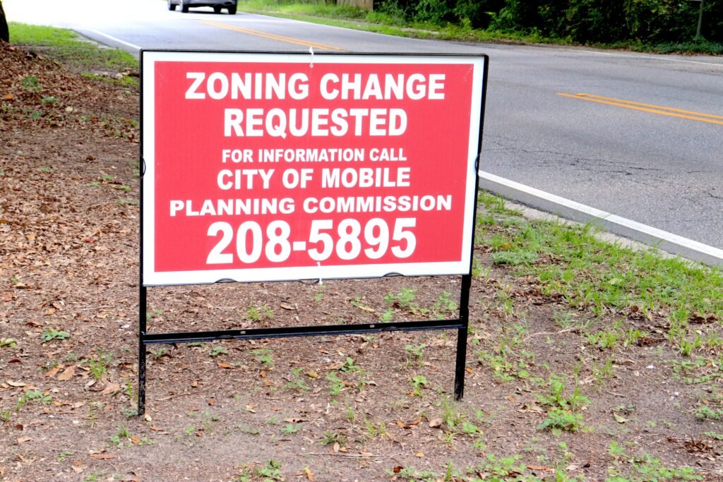 A red and white roadside public notice sign reading "Zoning Change Requested" with a phone number for the City of Mobile Planning Commission.