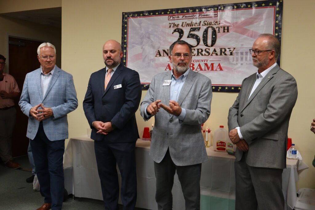 Four Republican officials standing together to address the audience. Acting & Vice Chairman Stuart Lang and candidate Chris Pringle speak to the crowd using hand gestures while standing in front of a white "United States 250th Anniversary Alabama" banner. Jugs of Milo's sweet tea sit on a refreshments table behind them.