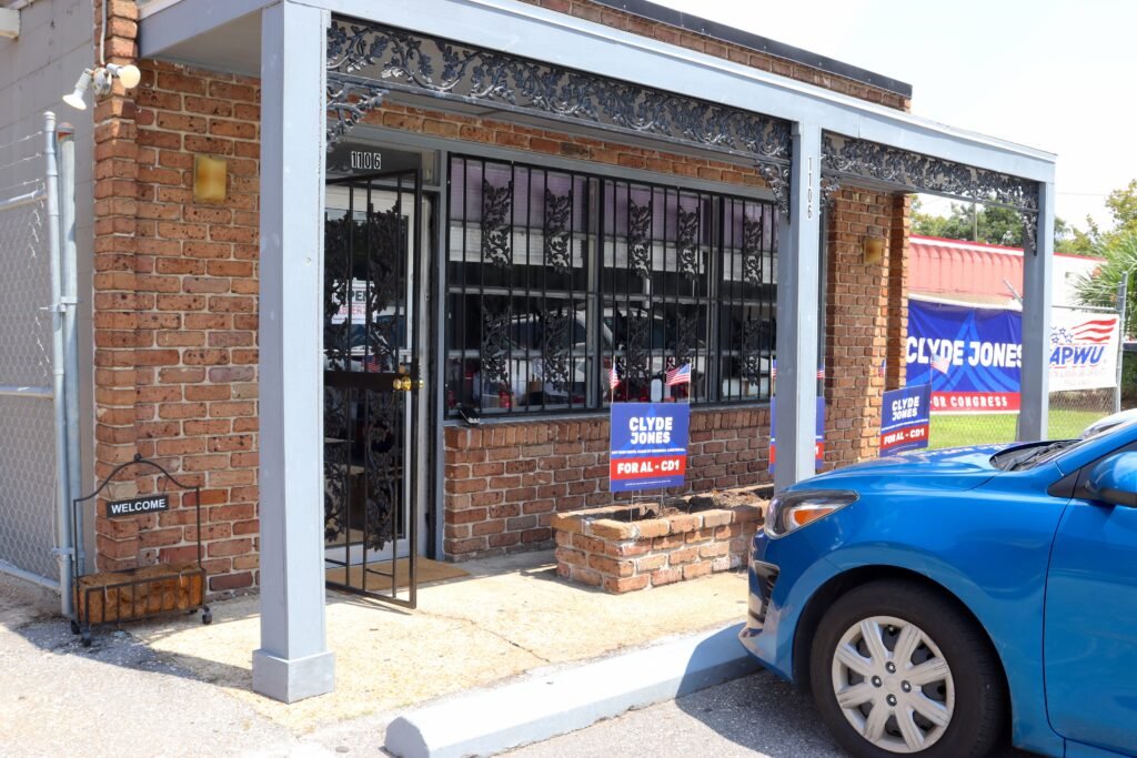 The brick exterior of the Clyde Jones for Congress headquarters at 1106 Spring Hill Avenue in Mobile, displaying blue yard signs for the AL-CD1 congressional race near a blue car in the foreground.