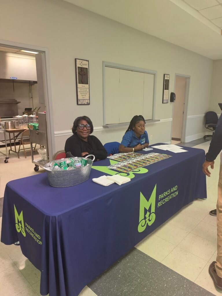 Two women sit behind a blue table cloth displaying the green Mobile Parks and Recreation logo.