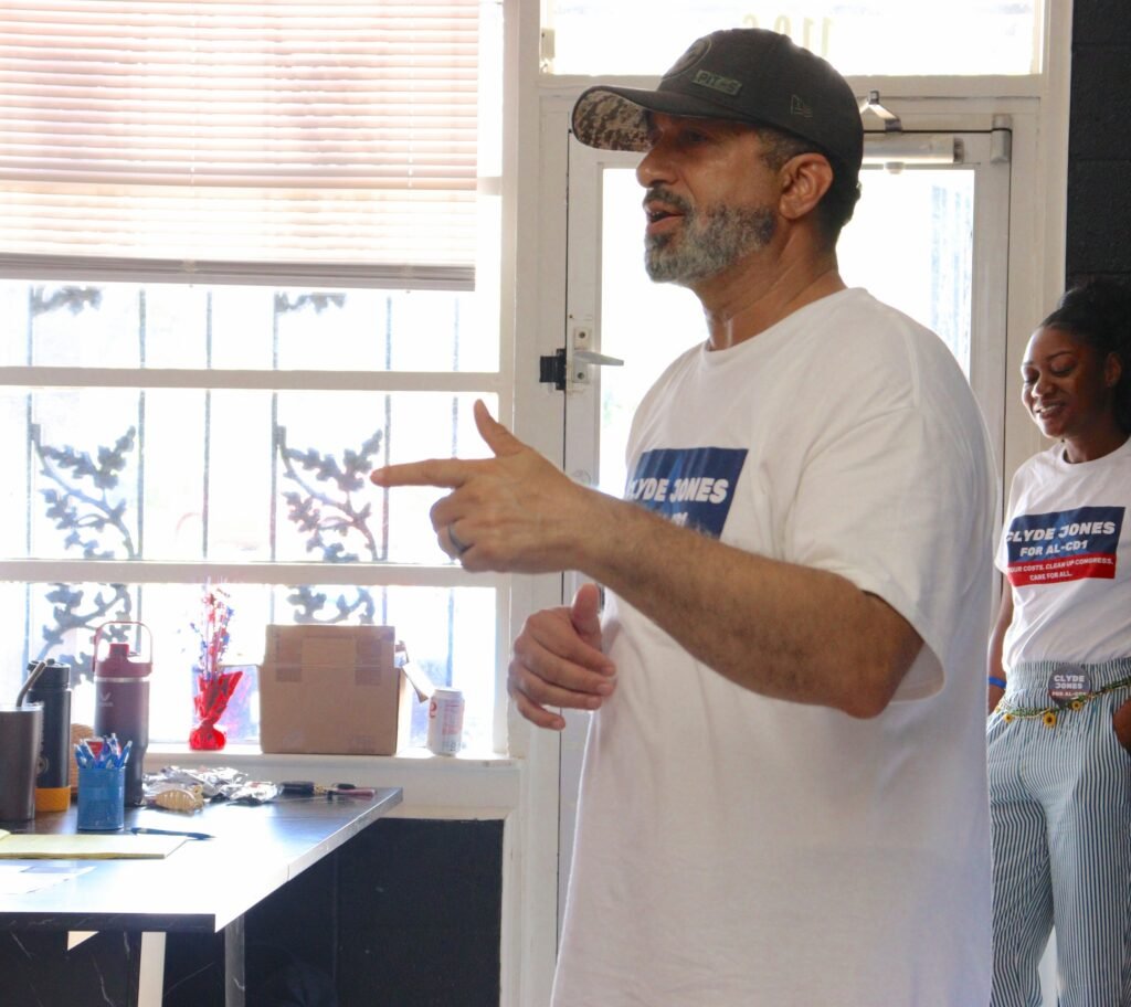 Candidate Clyde Jones wearing a campaign t-shirt and baseball cap while speaking and gesturing to a crowd inside the Mobile headquarters.
