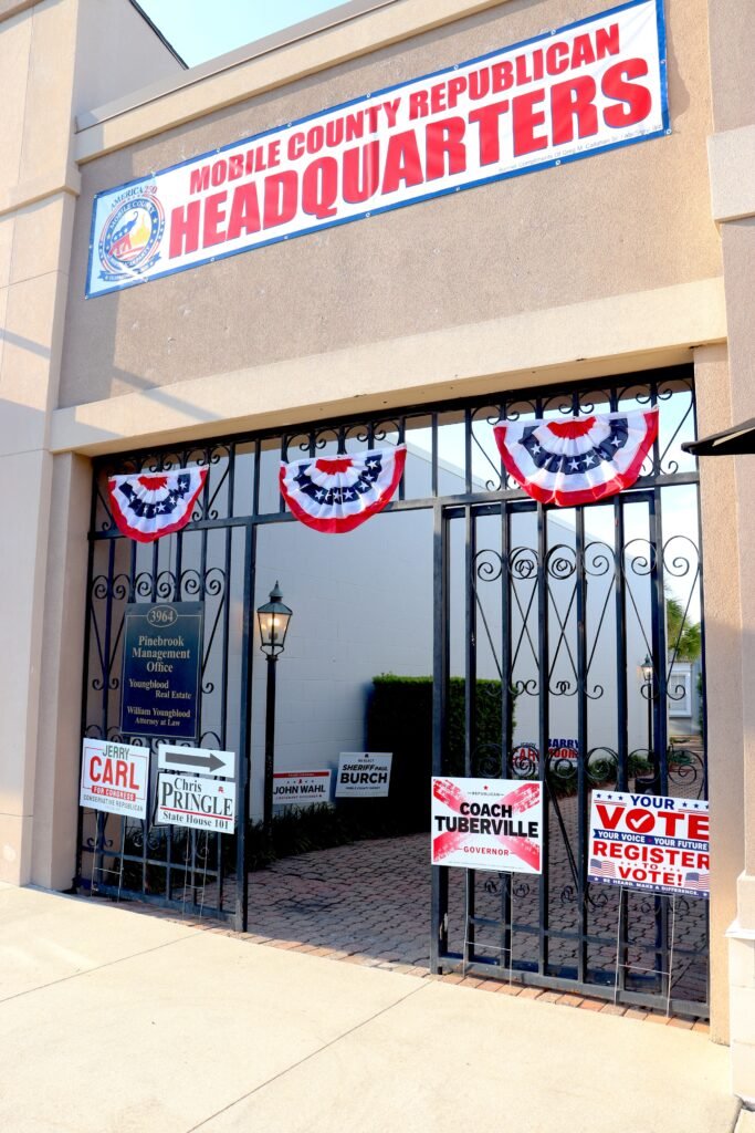 The exterior entrance of the shared Mobile County Republican Headquarters. A large printed banner over the arched walkway reads "Mobile County Republican Headquarters." The iron gates below are decorated with patriotic red, white, and blue bunting, alongside multiple campaign and voter registration signs.