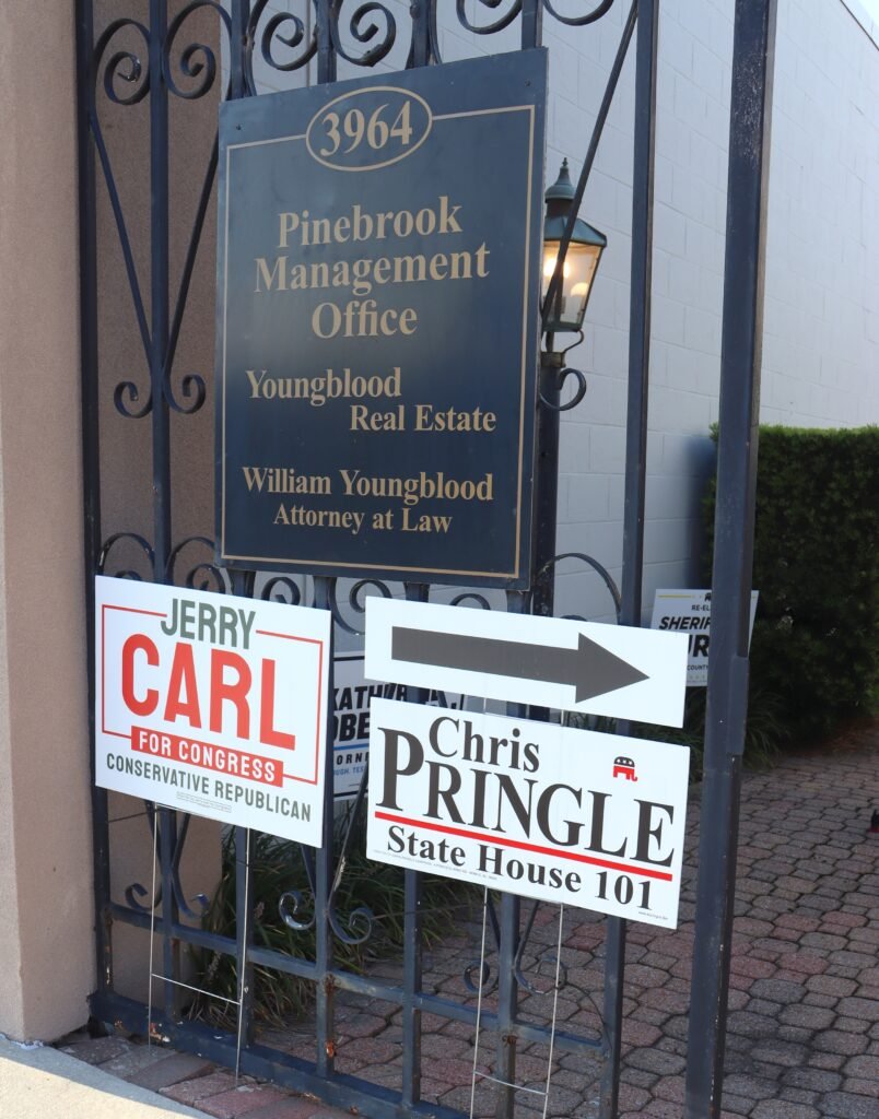 Campaign signs attached to an iron security gate. A dark plaque marks the "3964 Pinebrook Management Office." Below it, a white yard sign points visitors to the right for the Chris Pringle State House 101 office, alongside a sign supporting Jerry Carl for Congress.