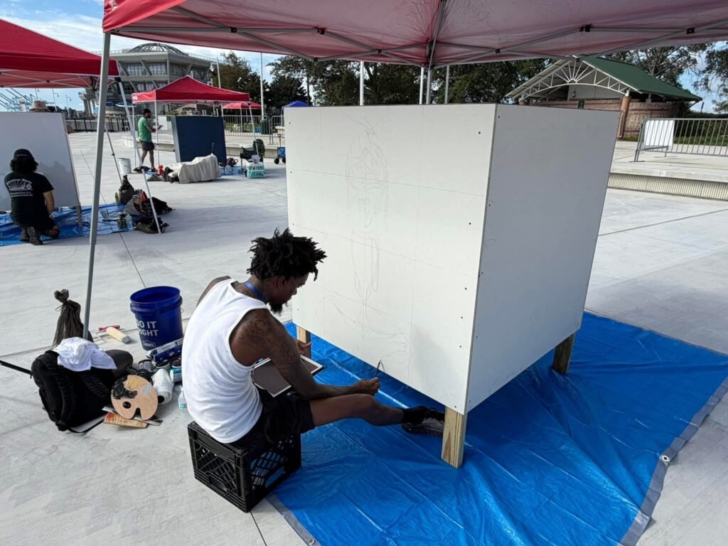 An artist sits on a crate sketching line work onto a large white marine-grade art cube under a red canopy tent, with other participants working at separate stations in the background.