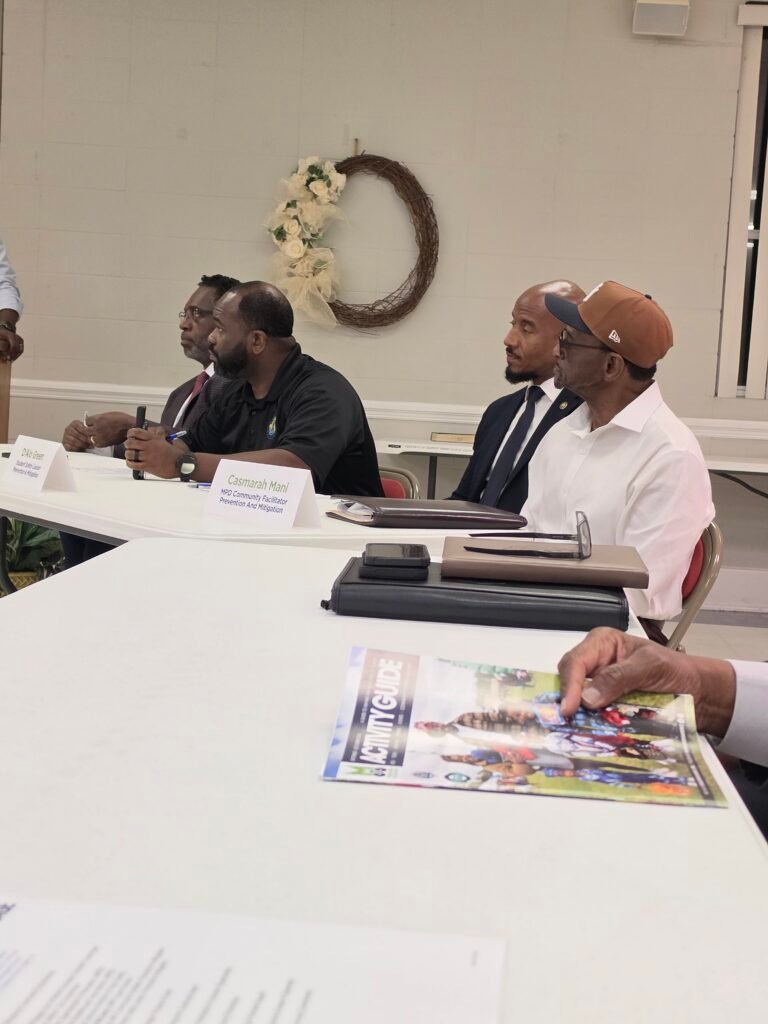Four men sit behind a white folding table with visible name cards including Casmarah Mani.