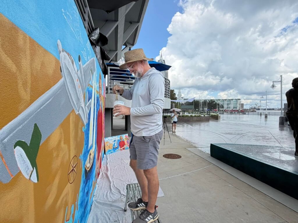 An artist wearing a straw hat and grey shirt paints a detailed mural featuring an airplane and a sailboat on an outdoor wall in downtown Mobile.