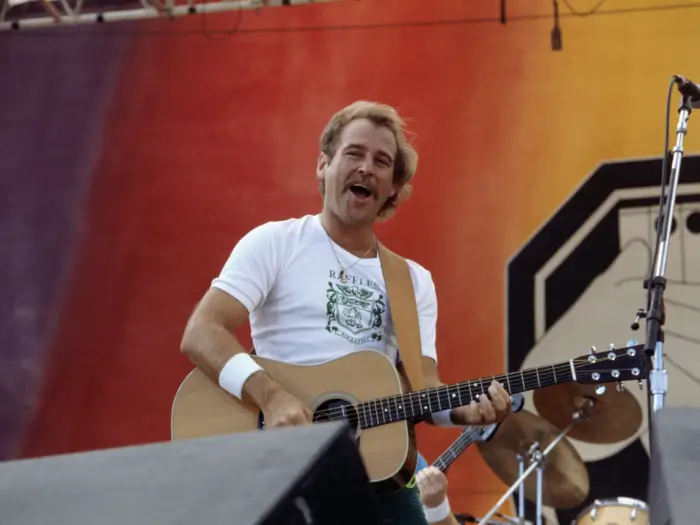 A vintage photo of a young Jimmy Buffett performing live on stage with an acoustic guitar.