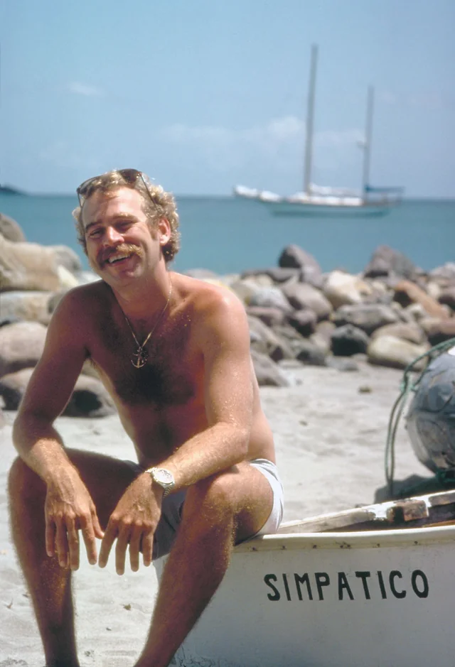 A young Jimmy Buffett smiling while sitting on a small boat named Simpatico on a beach with a sailboat in the background.