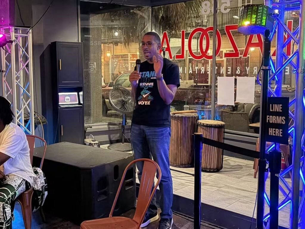 Stand Up Mobile representative Reggie Weaver speaking into a microphone on stage during the event at Kazoola in downtown Mobile, Alabama.