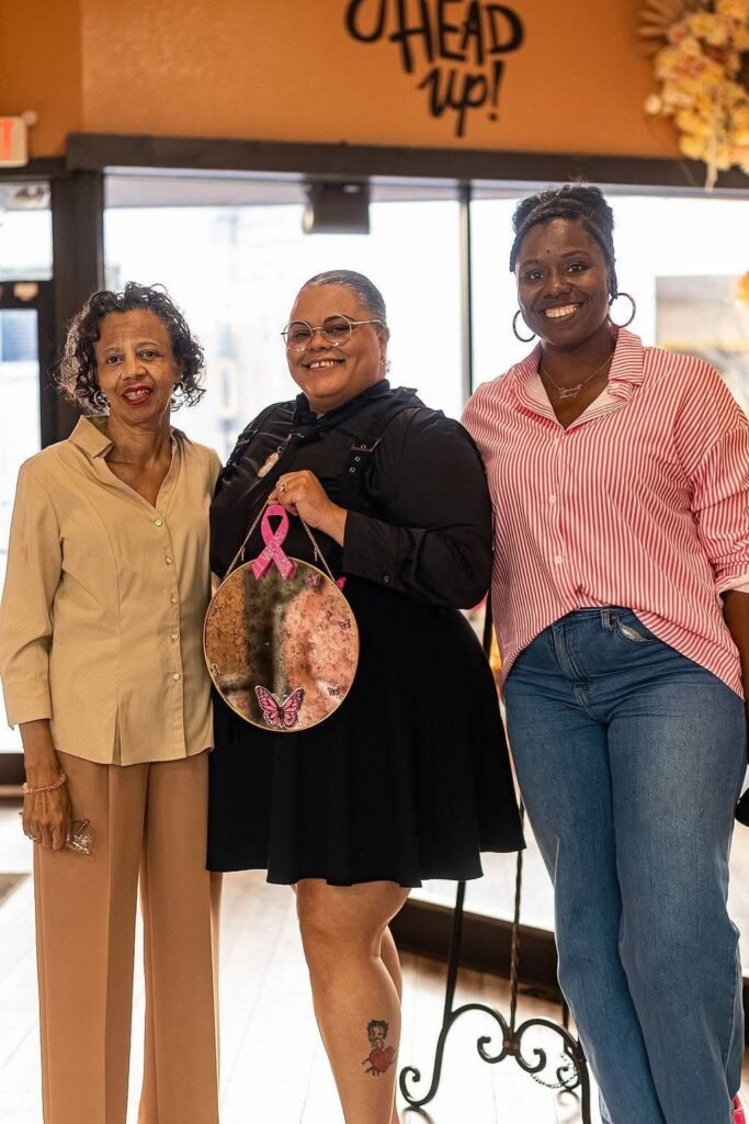 India Washington and local participants stand together inside Great Day Latte while holding a decorative circular mirror hanging from a pink breast cancer awareness ribbon.