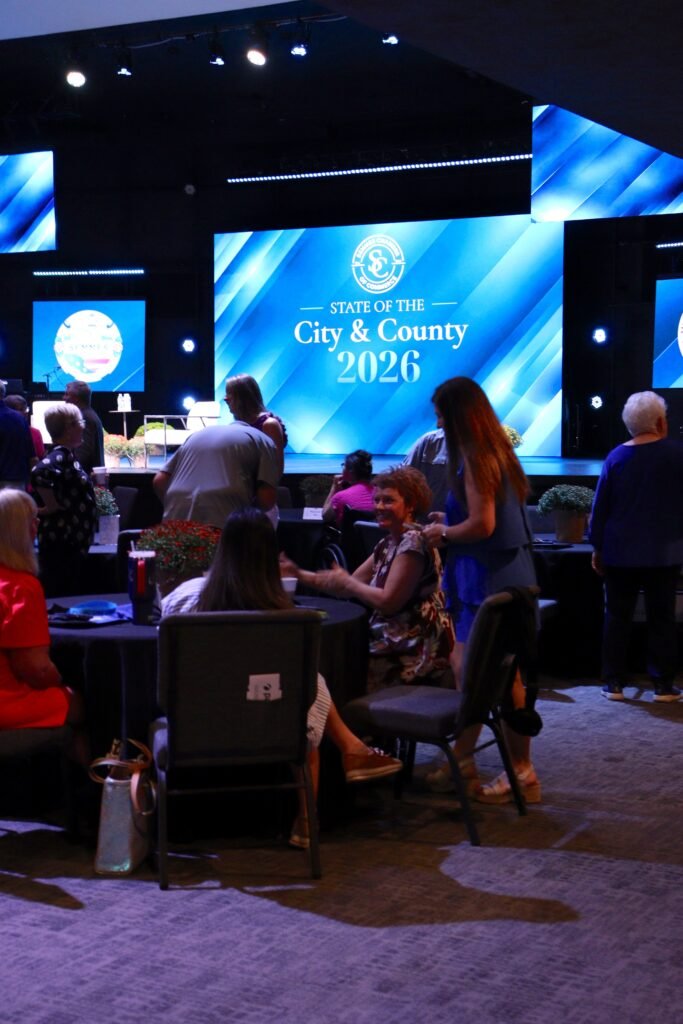 Attendees gather around round tables in the sanctuary of Pathway Church before the event begins. A large digital screen on the main stage displays the logo for the State of the City and County 2026.
