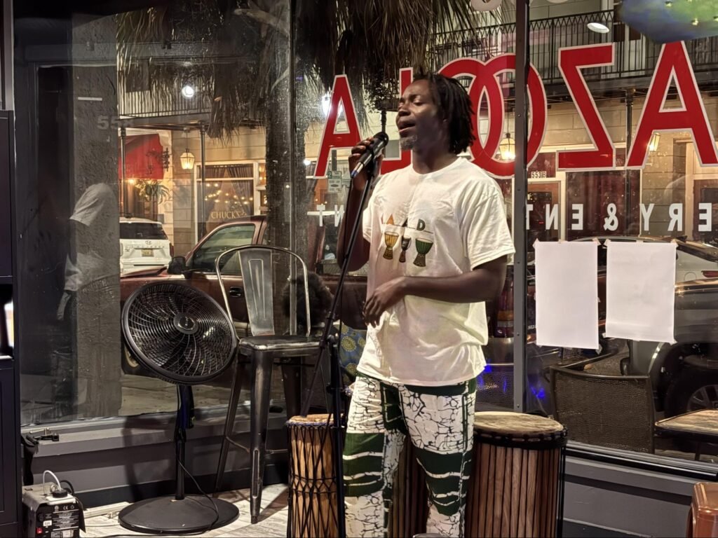 Benin artist Yewhe Yeton singing into a microphone on stage next to traditional drums at Kazoola in Mobile, Alabama.