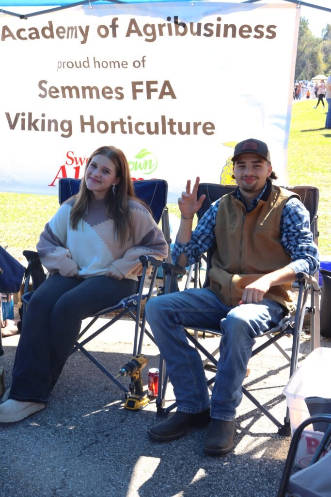 Two students sitting in lawn chairs under a banner for the Academy of Agribusiness, Semmes FFA, and Viking Horticulture at the festival.