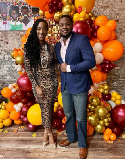 An indoor medium full shot of event host Alvin Jarreau Jr. and an attendee standing side-by-side in front of an elaborate balloon installation at the "FOR HER" event.
