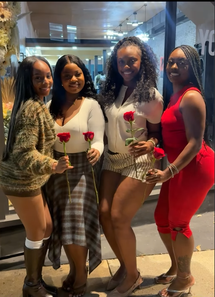 An outdoor medium full shot of four female attendees posing together outside the venue entrance at night. Each woman is smiling and holding a single red rose. From left to right: the first woman wears a green patterned sweater dress with dark knee-high boots; the second wears a long-sleeved white top and a plaid skirt; the third wears a white short-sleeved top and striped shorts; the fourth wears a sleeveless red jumpsuit and metallic sandals. The window of the brightly lit venue is visible behind them.