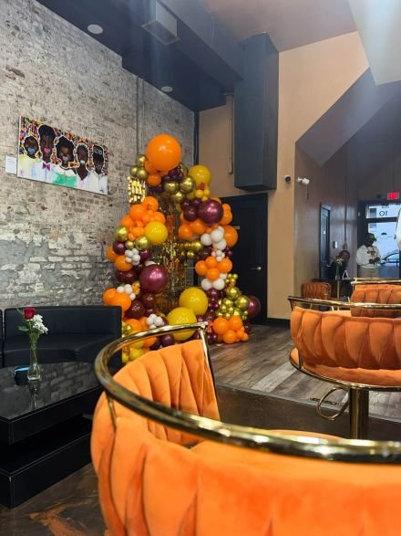 An interior view looking through the seating area of Great Day Latte during the event setup. In the immediate foreground, the backs of three curved, orange velvet stools with gold-colored frames are visible. The middle ground features a black coffee table with a small floral arrangement next to a dark gray lounge sofa. In the background, a large decorative balloon arch in orange, gold, and maroon stands against an exposed brick wall next to framed artwork.