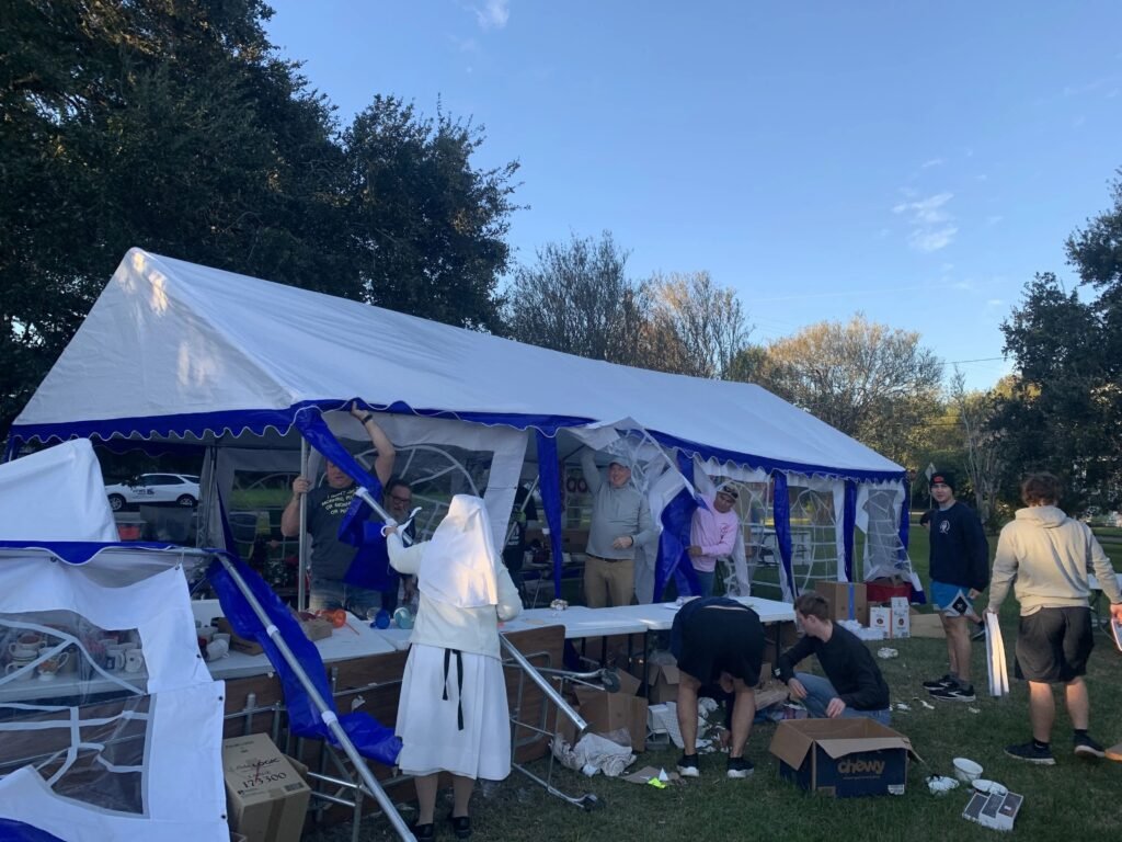 A Catholic nun and several people work to reconstruct a large, wind-damaged white and blue event tent on a grassy lawn the morning after a storm. Long folding tables, cardboard boxes, and debris are scattered across the grass as they lift the canvas and secure the metal frame.